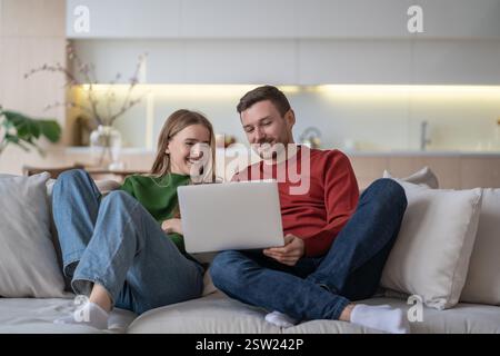 Happy loving couple smiling watching fun movie using laptop comfortable sitting on couch at home. Stock Photo