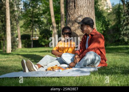 Happy relaxed loving black couple sits on grass chatting, enjoying picnic in park resting together. Stock Photo