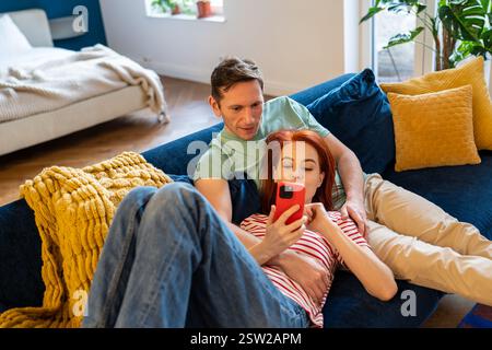Loving couple resting on couch using smart phone apps. Wife showing interesting website to husband Stock Photo
