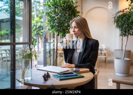 Thoughtful businesswoman takes moment to drink coffee after working on laptop in cafe. Tea break. Stock Photo