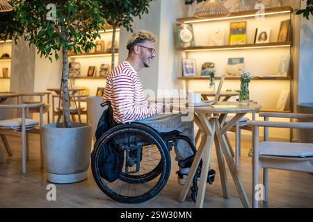 Happy pleased man on wheelchair in cafe using laptop for communication, video call, online chatting. Stock Photo