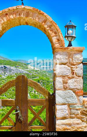 Megali Panagia Monastery entrance, Greece. A stone arch with lanterns and a wooden gate opens to panoramic views of lush hills and a bright blue sky. Stock Photo