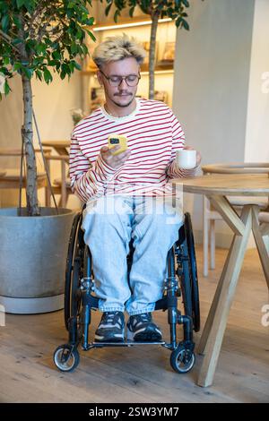 Thoughtful dreamy disabled man in wheelchair drinking coffee at cafe table, using mobile phone Stock Photo