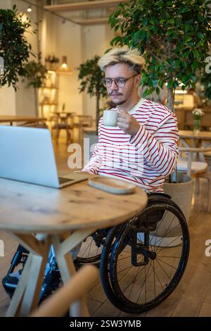 Focused guy freelancer with disabilities in wheelchair working online using laptop sits in cozy cafe Stock Photo
