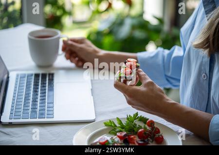Peaceful woman at table in morning holding sandwich having breakfast using laptop computer. Stock Photo