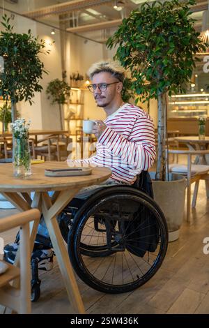 Thoughtful dreamy disabled man in wheelchair drinking coffee at cafe table, waiting for meeting Stock Photo