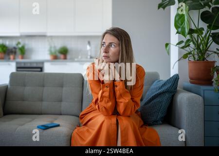 Worried stressed woman feel tension sitting on sofa looking aside waiting call on cellphone at home. Stock Photo