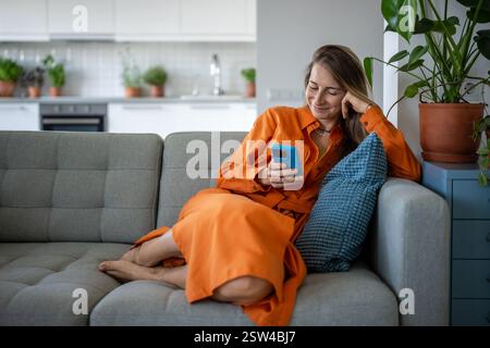 Happy woman using cellphone at home sofa. Pleased relaxed female have internet fun, chatting online. Stock Photo