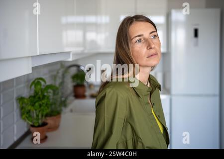 Pensive serious thoughtful middle aged woman sadly looking has bad mood standing on kitchen at home. Stock Photo