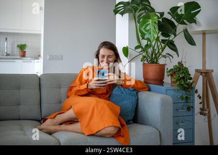 Happy woman using cellphone at home sofa. Pleased relaxed female have internet fun, chatting online. Stock Photo