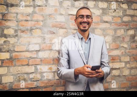 Portrait of happy young businessman who is messaging on phone. Young businessman standing in the front of brick wall and using smartphone. Stock Photo