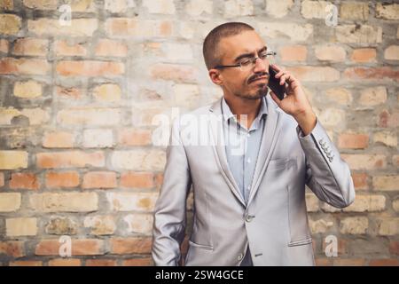 Portrait of distraught businessman who is talking on phone. Young businessman standing in the front of brick wall and using mobile phone. Stock Photo