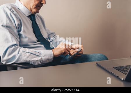 Senior businessman multitasking with smartphone and laptop at office desk. Close-up of hands using a mobile device for communication and productivity. Stock Photo
