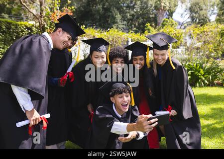 Graduating diverse students celebrating in garden, taking selfie with joyful smiles and diplomas. Graduation, celebration, diverse friends, achievemen Stock Photo