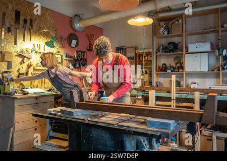Artisan collaborating with apprentice assistant, constructs bed frame from solid wood in workshop Stock Photo