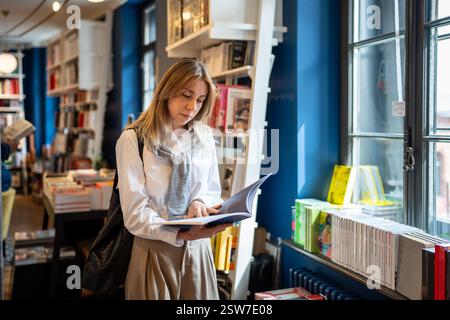 Focused thoughtful middle aged woman reading book standing shopping in modern bookstore near window Stock Photo