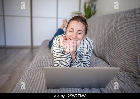 Happy middle aged woman have fun using laptop smiling lounging on couch watching interesting movie. Stock Photo