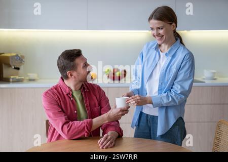 Happy young couple having breakfast, wife serves tea to husband in kitchen, love and devotion Stock Photo