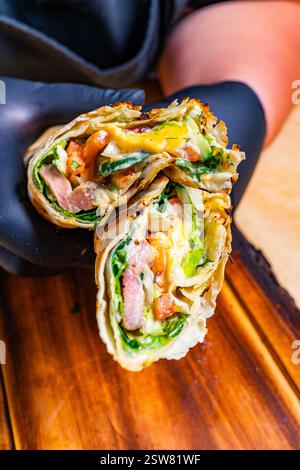 A person wearing black gloves holds a freshly made wrap filled with greens, tomatoes, cheese, and meat. The wrap is cut in half to reveal its colorful Stock Photo