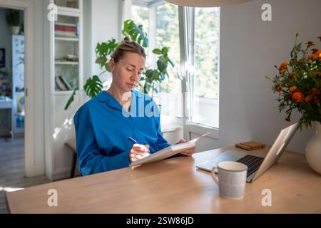 Focused on diary woman planning day in morning sitting at home with mug coffee, laptop writing notes Stock Photo