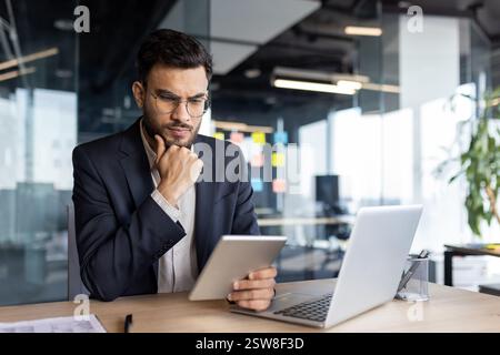Concentrated young entrepreneur in suit working with a tablet at desk in the office. Solving business problems Stock Photo