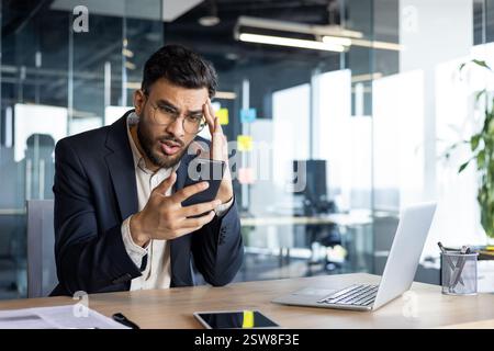 Upset and disappointed man with phone in hands inside office at workplace, businessman reading bad news Stock Photo