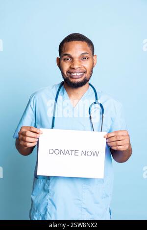 African american nurse advertising the message to donate blood, holding a paper and urging people to help in studio. Medical assistant in workwear standing against blue background. Stock Photo