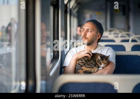 Relaxed man commuting in electric train with cat comfortably sitting in hands, traveling with feline Stock Photo