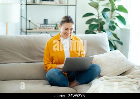 Smiling young caucasian woman in a yellow shirt sitting cross-legged on a cozy sofa, using a laptop in a bright modern living room. Concept of remote work, freelance, and online communication Stock Photo