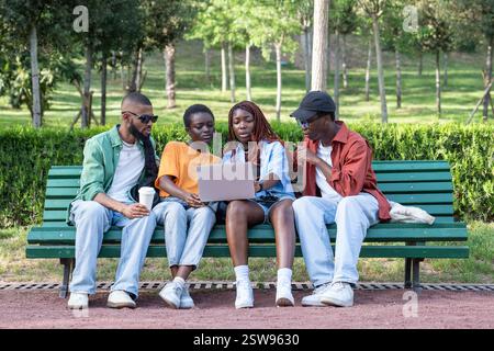 Pleased relaxed group of black students sitting on bench together looking to laptop on girls knees Stock Photo