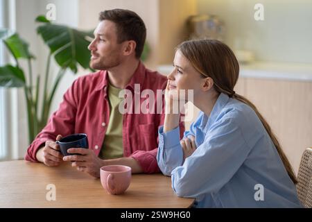 Smiling young couple staring into the distance while having tea and breakfast together in kitchen Stock Photo