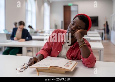 Sleepy lazy African American man falls asleep sits in library with head resting on desk with books Stock Photo