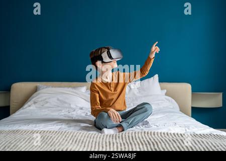 Child boy wearing virtual reality headset sitting on bed at home exploring metaverse. VR concept Stock Photo