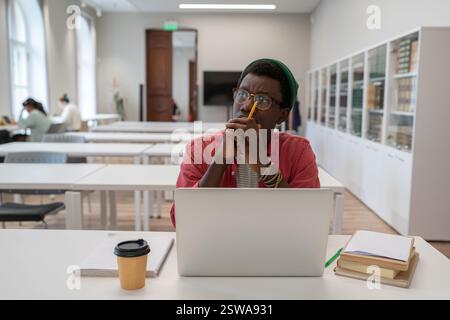 Thoughtful african american nerd man student studying in library using laptop. Stock Photo