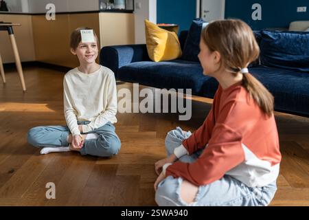 Smiling teen sister and child brother playing who am i game sitting on floor at home in living room. Stock Photo