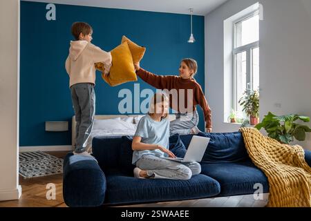 Playful european siblings boy and girl pillow fighting distracting busy mom relaxing on sofa at home Stock Photo