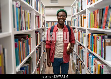 Smiling african american man student holds book in university library between bookshelves. Stock Photo