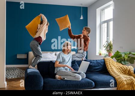 Naughty children fun playing pillow fight near busy smiling mother with laptop sitting on home sofa Stock Photo