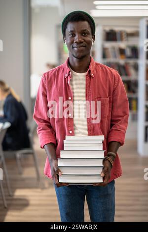 Portrait of happy African American student guy with pile of books in library Stock Photo