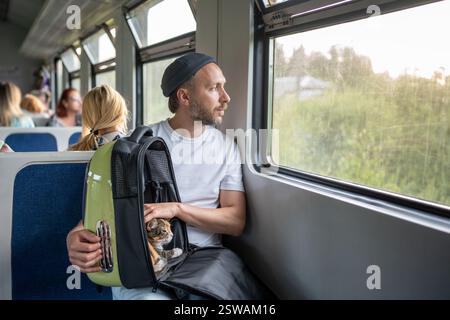 Thoughtful man owner calming stroking cat in pet backpack while look out window on train. Pet safety Stock Photo
