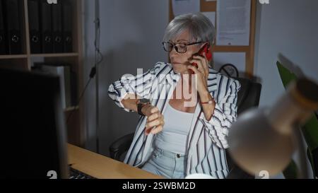 Mature woman in an office checking her watch while talking on a phone. Stock Photo