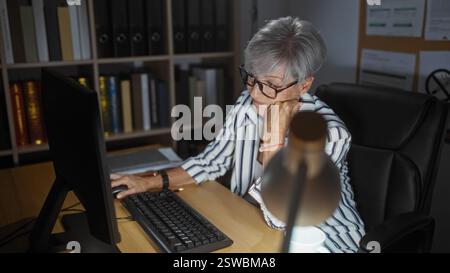 Mature woman working on a computer in an office, wearing glasses and a striped shirt, surrounded by books and binders, looking focused and engaged in Stock Photo