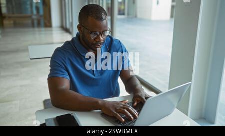 Young, african, american, man working indoors in a modern office setting, focusing intently on his laptop with glasses and a blue shirt Stock Photo