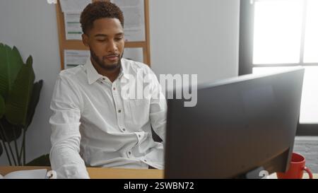 Young man working at a desk in an office setting, focused on his computer with a plant in the background Stock Photo