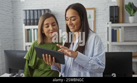 Two women working together in an office, one showing something on a tablet to the other, both smiling and appearing engaged in their workplace discuss Stock Photo