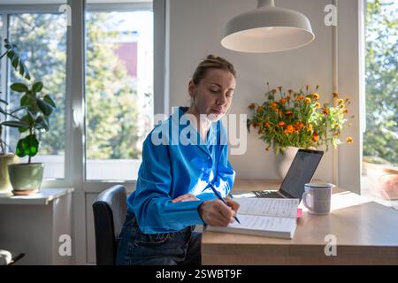 Focused woman comfortable works at home by writing notes in day planner sits at table with laptop. Stock Photo
