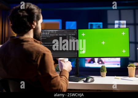 Programmer drinking hot beverage and using green screen PC for futureproofing company network from downtimes and unexpected system failures. Admin with coffee mug in front of mockup computer Stock Photo