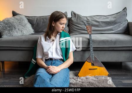 Thoughtful female musician sit on floor, observe balalaika. Challenge of mastering traditional music Stock Photo