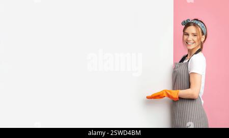 Young Housewife In Rubber Gloves Pointing At Empty White Advertisement Board Stock Photo