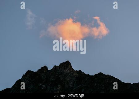 Fiery cloud in the evening over the dark silhouette of sharp mountains. Sky at sunset. Pink bright red cloud in the evening sky. Stock Photo